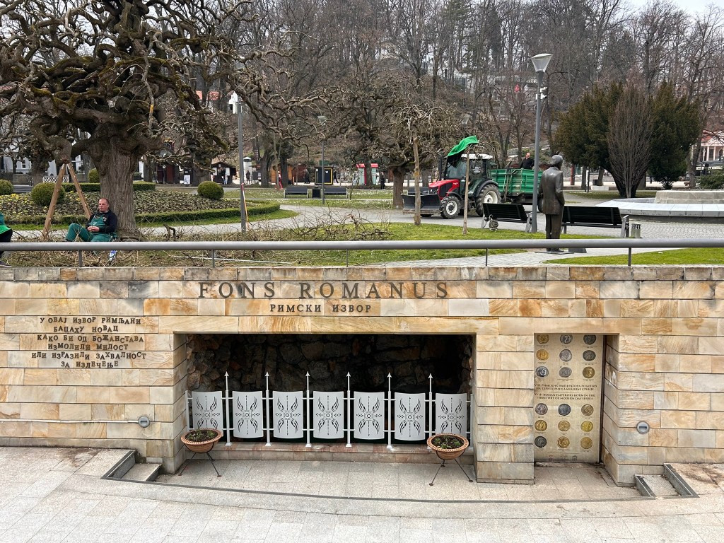 Exterior view of Fons Romanus, a historical site with a stone entrance and decorative iron gates, set in a park with manicured greenery and surrounding trees.