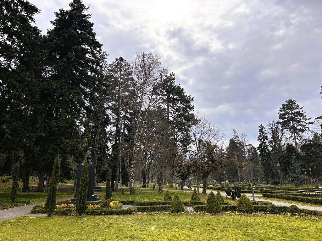 A serene park scene featuring tall trees, neatly manicured hedges, and a statue amidst a cloudy sky.