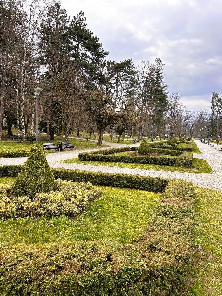 A landscaped park featuring neatly trimmed hedges, ornamental bushes, and stone pathways, with benches and tall trees lining the area under a cloudy sky.