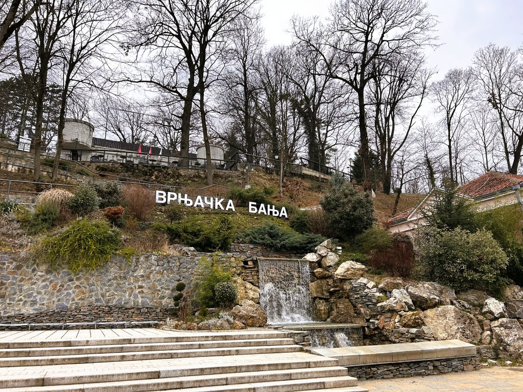 A scenic view of a hillside in Vrnjačka Banja, featuring a stone wall, greenery, and a small waterfall, with the name 'ВРЊАЧКА БАЊА' prominently displayed in white letters.