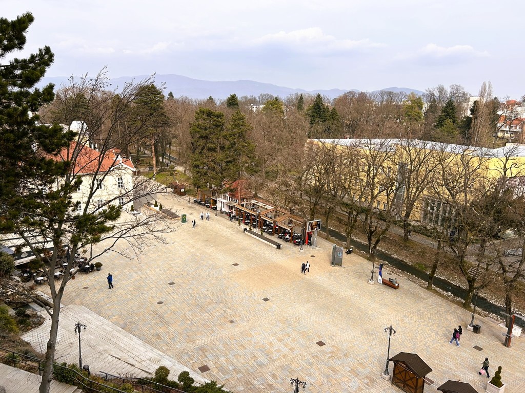 A panoramic view of a spacious outdoor area lined with trees, featuring a small café and people walking. The scene is set against a backdrop of mountains under a cloudy sky.