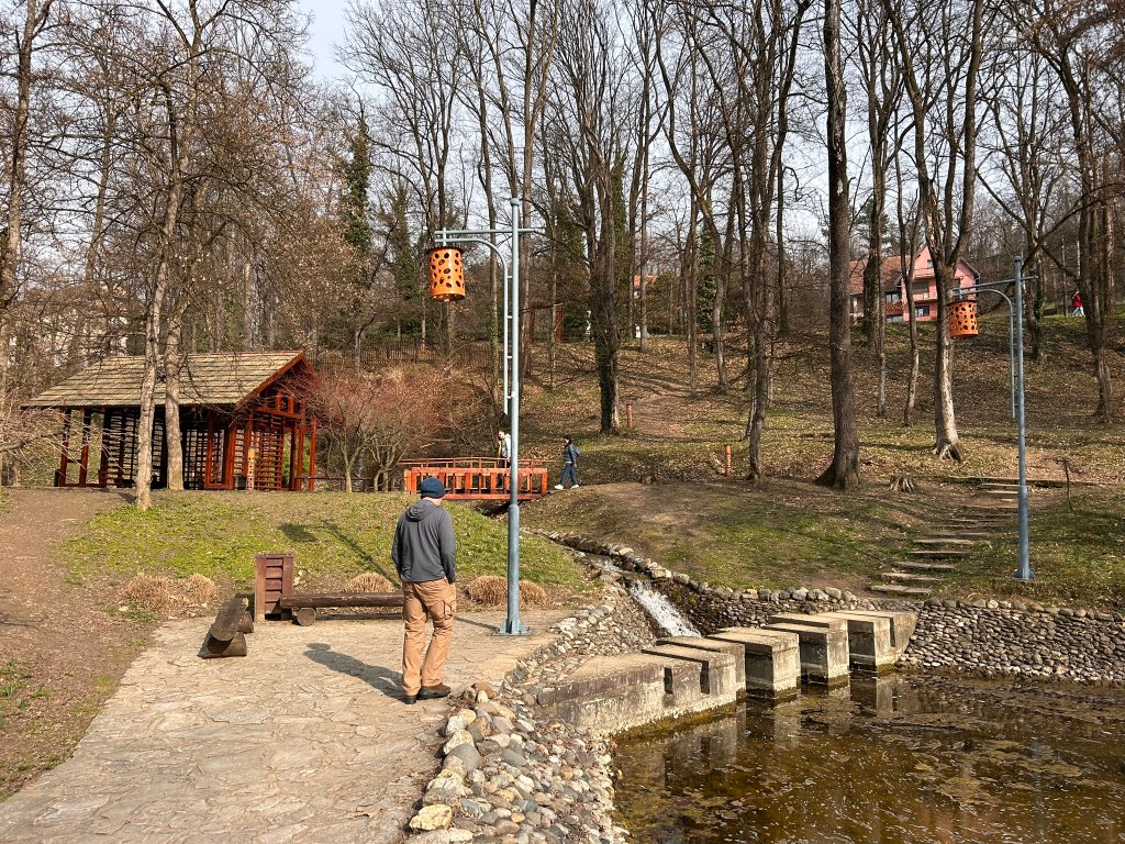 A man walks along a stone path by a pond in a park with trees and a wooden structure in the background.