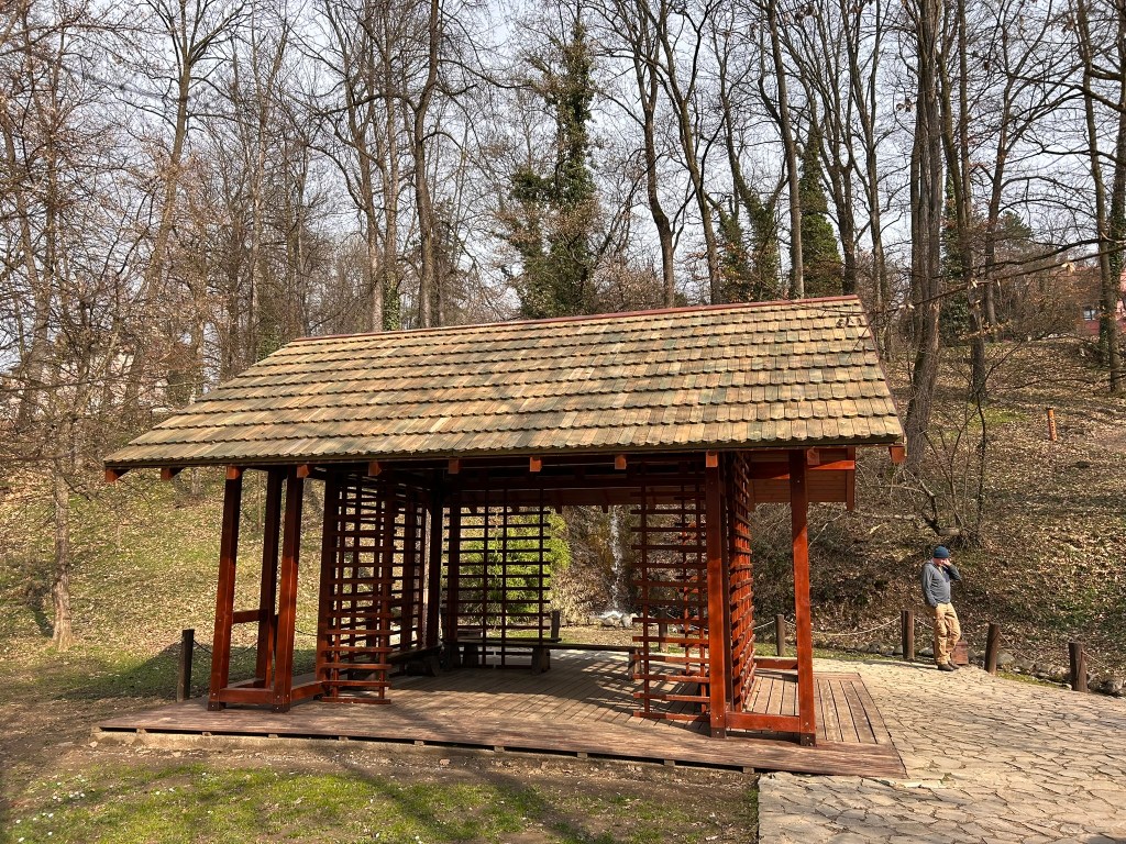 A wooden gazebo with a tiled roof situated in a forested area, surrounded by bare trees and a stone pathway, with a person standing nearby.