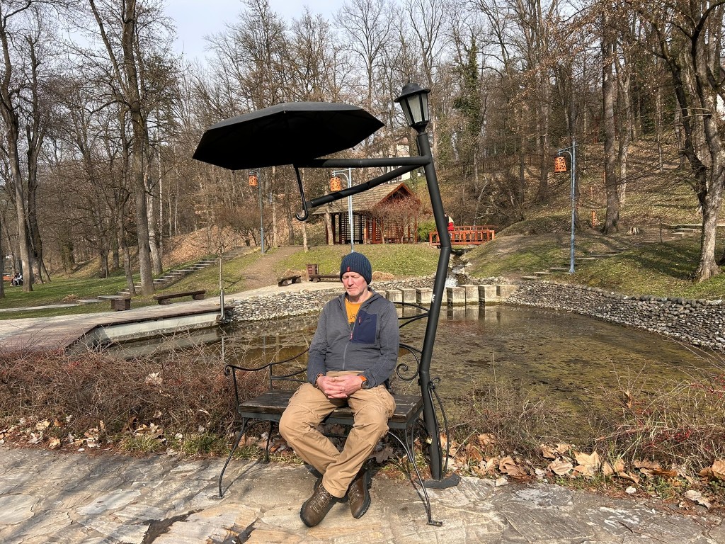 A man sitting on a black bench with a parasol in a park setting near a small pond, surrounded by bare trees and pathways.
