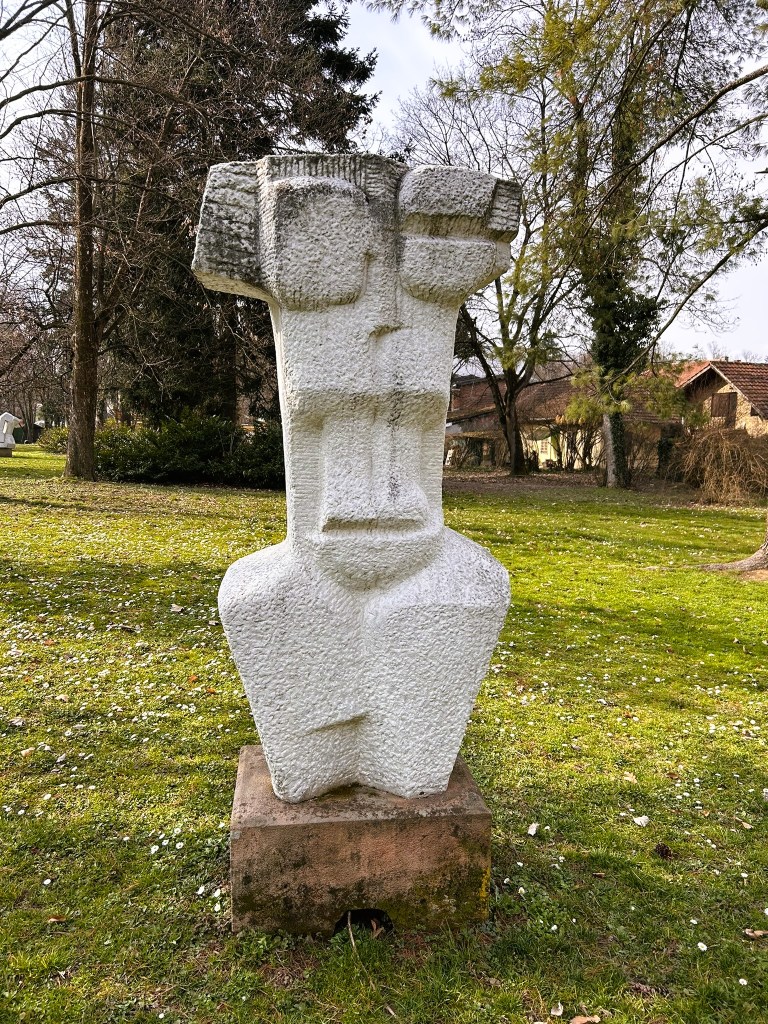 A white stone sculpture resembling a stylised human figure, set on a stone base, surrounded by greenery in a park.