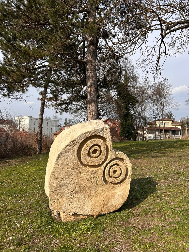 A carved stone sculpture featuring two circular designs, set on a grassy area with trees in the background.
