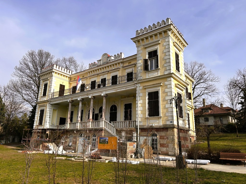 A partially renovated historic mansion with a yellow facade, featuring intricate architectural details, balconies, and a Serbian flag. Surrounding trees are bare, indicating early spring, with construction materials visible in the foreground.