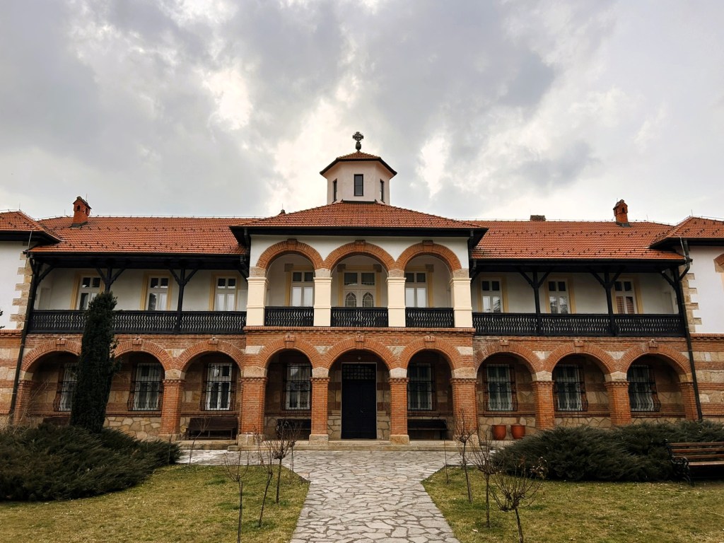 Facade of a historic building featuring arched colonnades and a central dome, surrounded by a grassy courtyard under a cloudy sky.