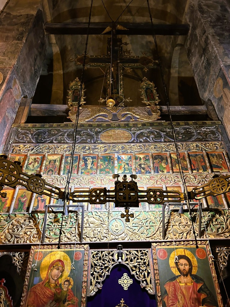 Interior view of a church altar featuring intricate wooden carvings, religious icons, and a large decorative cross mounted above a richly painted backdrop.