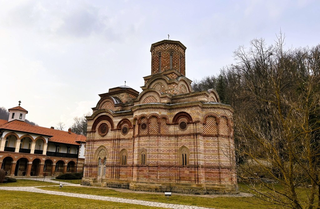 A historic brick church with intricate architectural details, surrounded by greenery and a tree, set against a cloudy sky.