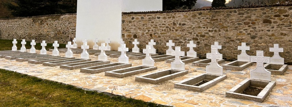 A row of white crosses marking graves in a cemetery, set against a stone wall.