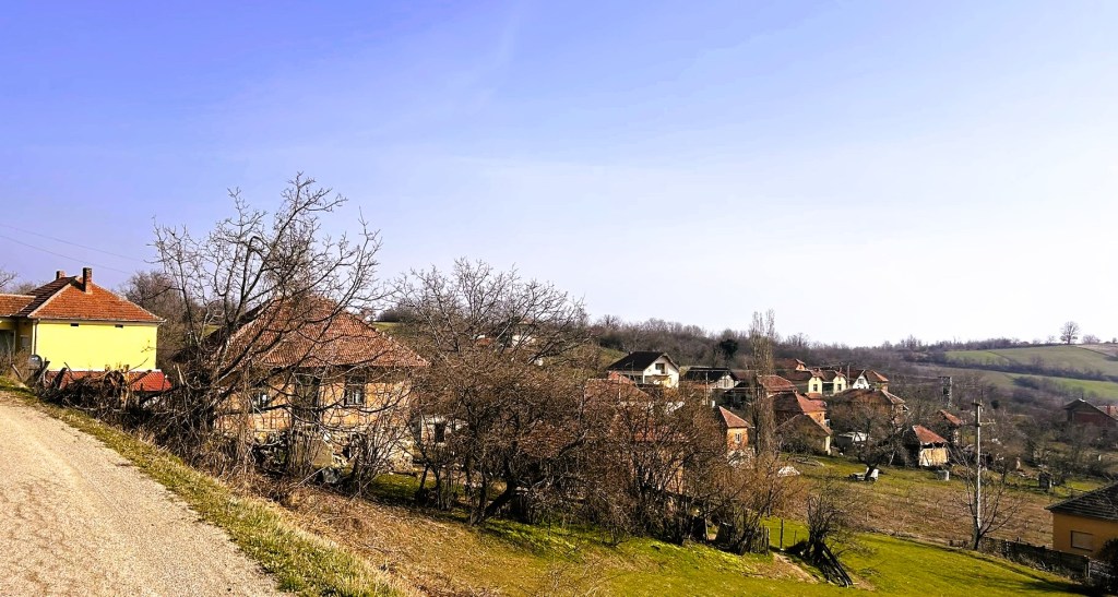 A rural landscape featuring a collection of houses with red and yellow rooftops, surrounded by bare trees and rolling hills under a clear blue sky.