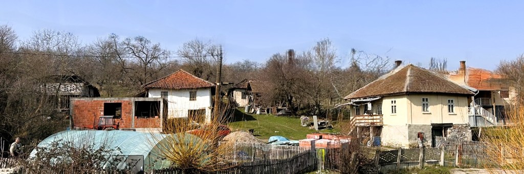 A rural landscape featuring several houses, including a partially constructed building, surrounded by sparse trees and greenery under a clear blue sky.