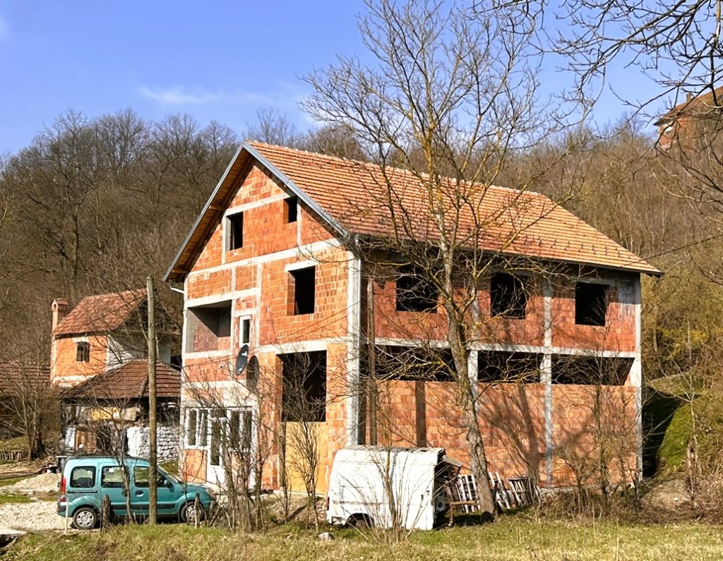 A partially constructed brick house with a tiled roof, surrounded by trees and a few vehicles in a rural setting.