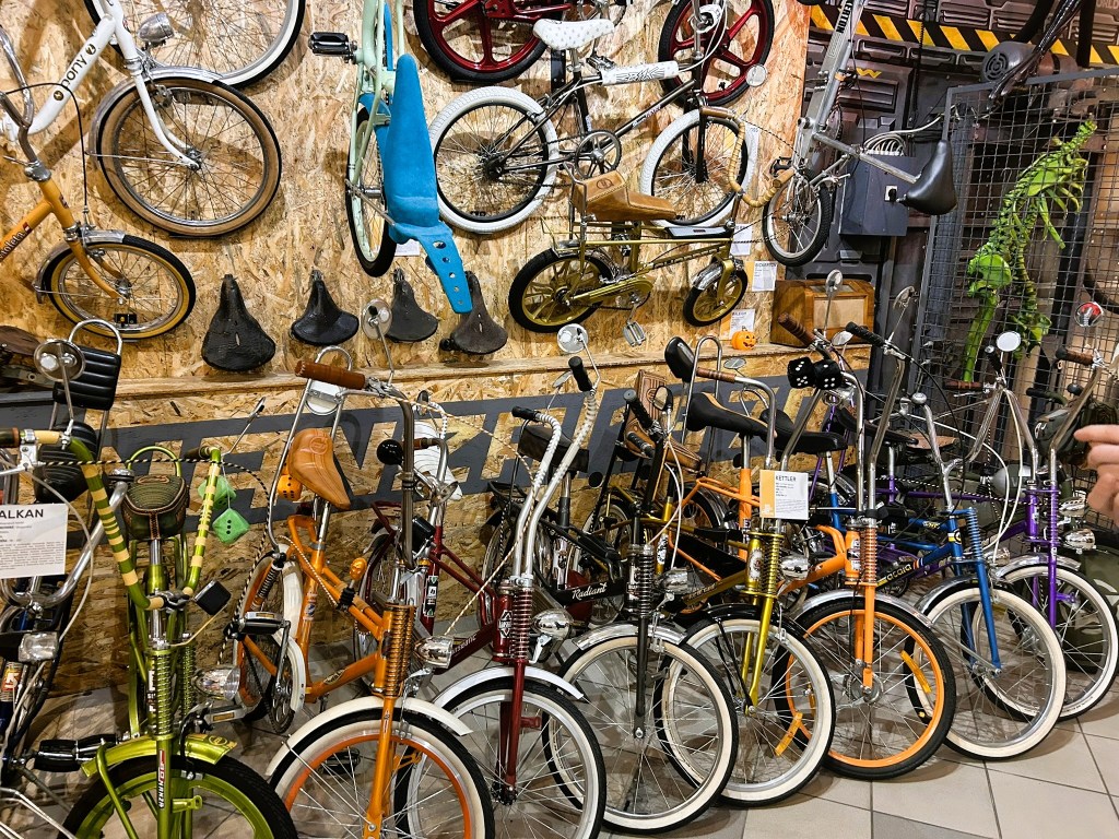 Display of vintage and retro bicycles in various colours, arranged against a wooden wall with additional bike parts and accessories hanging above.