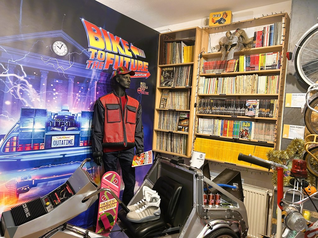 Display featuring a mannequin in a red and black outfit, standing next to a futuristic car and various items from the 'Back to the Future' franchise, with a bookshelf filled with comic books and records in the background.