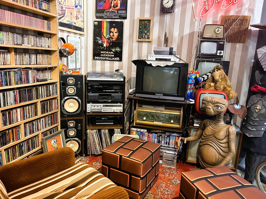 A vintage-style room corner featuring a collection of music CDs and cassettes on shelves, a retro audio system, a large TV set, and several nostalgic items, including a figurine of E.T. and plush toys. The room has a warm, inviting atmosphere with two patterned chairs and a variety of decorative posters on the walls.