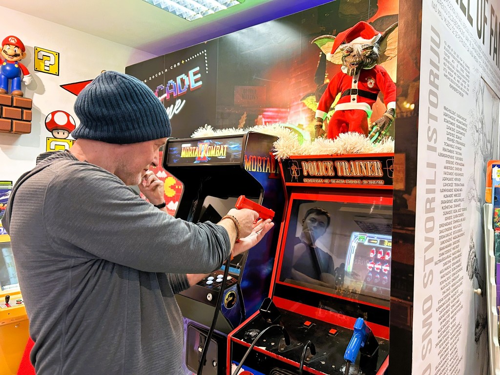 A person in a beanie playing an arcade game titled 'Police Trainer' in a gaming arcade, with a festive gremlin figure dressed as Santa in the background.