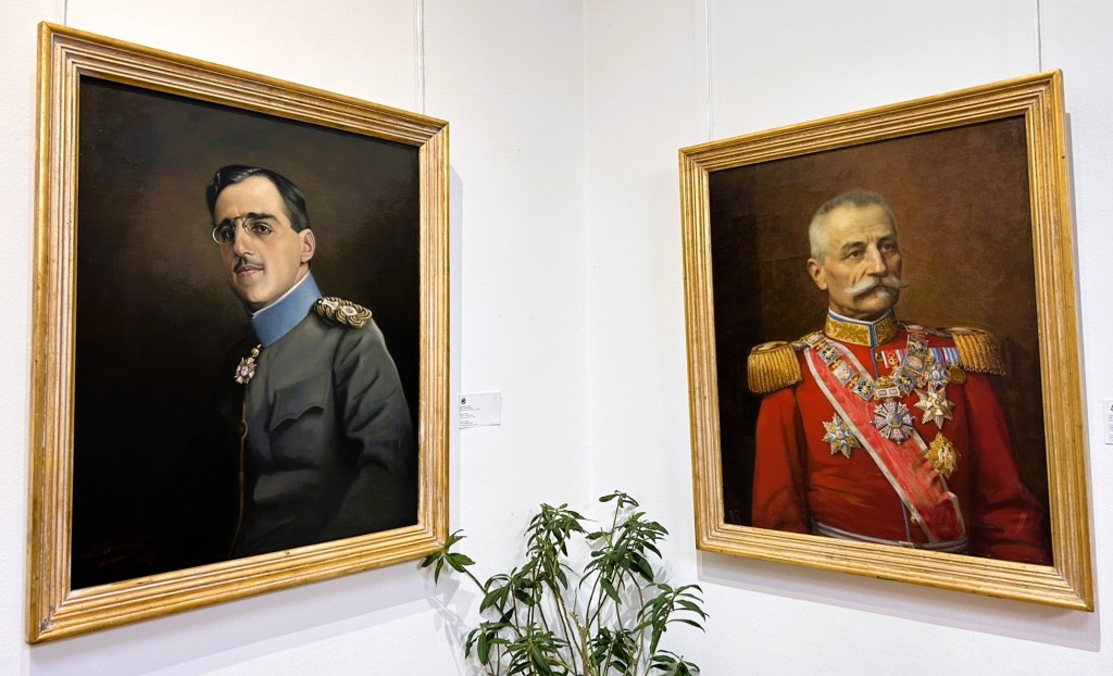 Two framed portraits of military figures displayed on a wall, one in a grey uniform with glasses, the other in a red uniform adorned with medals.