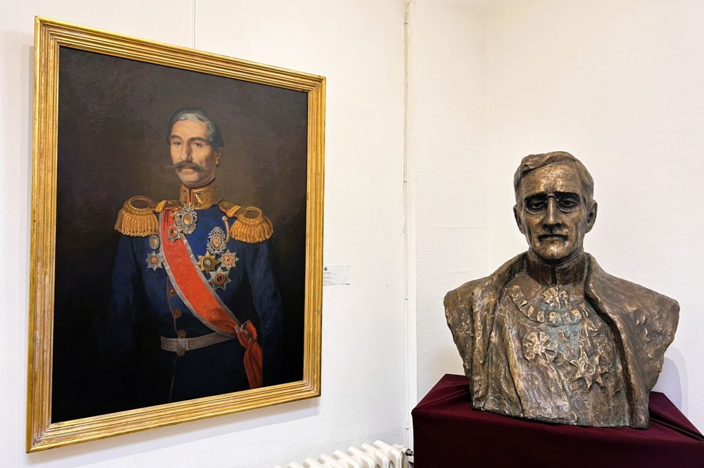 A portrait of a man in a military uniform with medals and decorations, framed in gold, displayed on the left. Beside it, a bronze bust of a man wearing a similar uniform, shown on a maroon cloth.