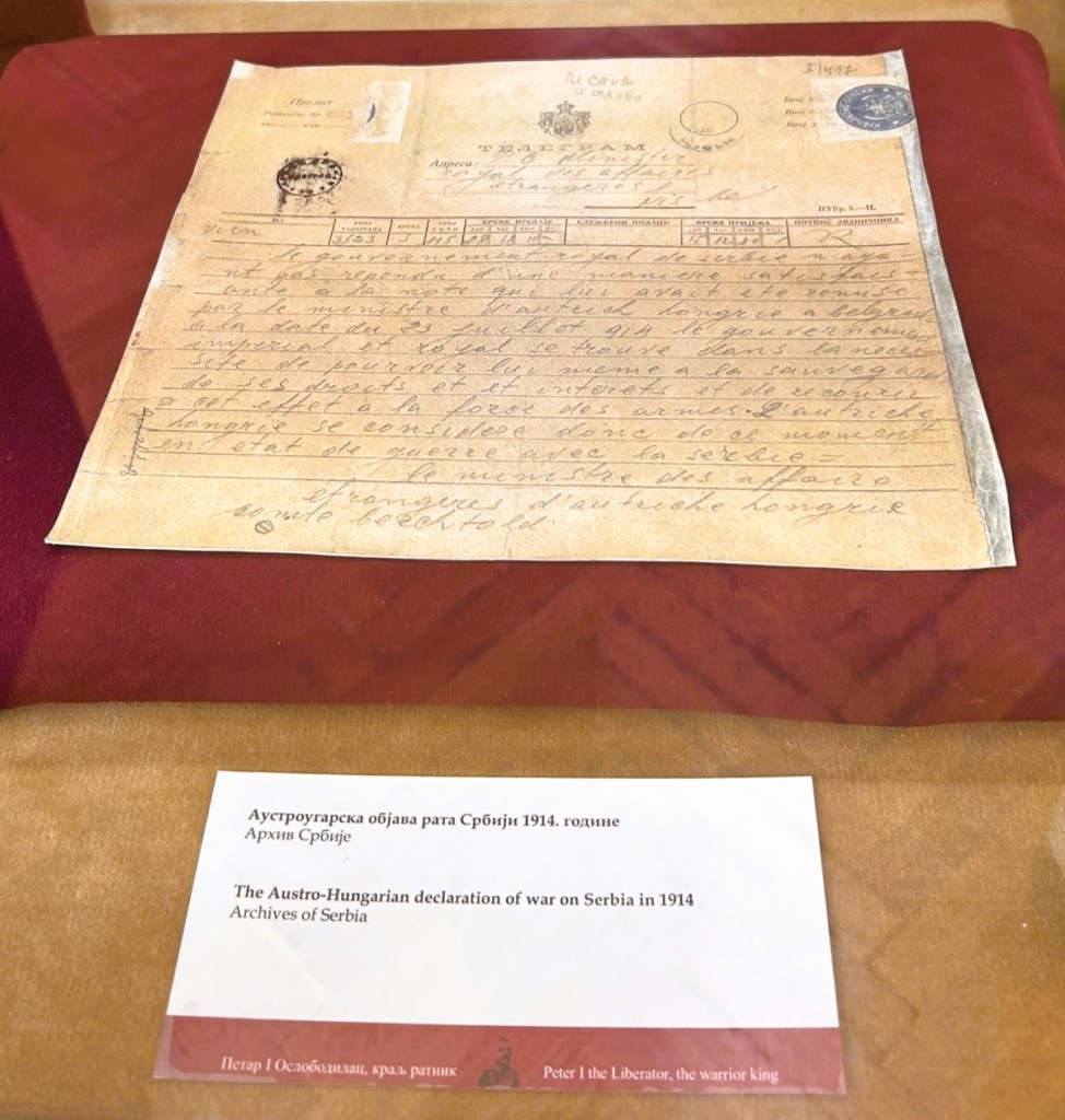 A historical document displayed on a red cushion, featuring the Austro-Hungarian declaration of war on Serbia from 1914. The document includes handwritten text and stamps, accompanied by a labelled explanation in both Serbian and English.