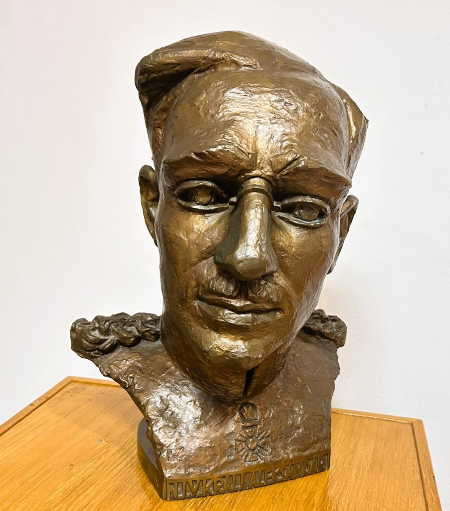 Bronze bust of a man with a prominent nose and serious expression, featuring a braided detail at the neck and an insignia on the chest, displayed on a wooden surface.