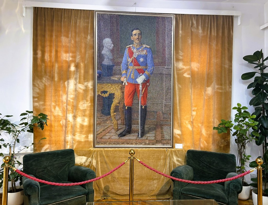 A portrait of a man in military uniform, standing beside an ornate table in a room with golden curtains and green armchairs, framed by a rope barrier and potted plants.