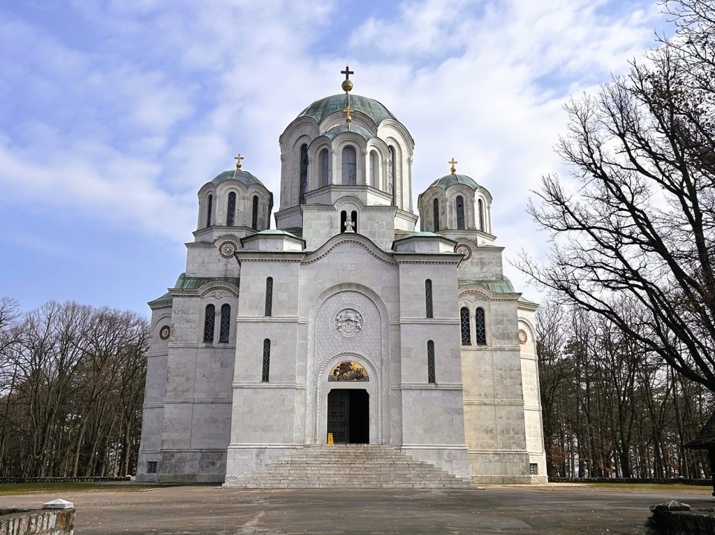 A grand white stone church with three domes and a large entrance, surrounded by trees and a cloudy sky.