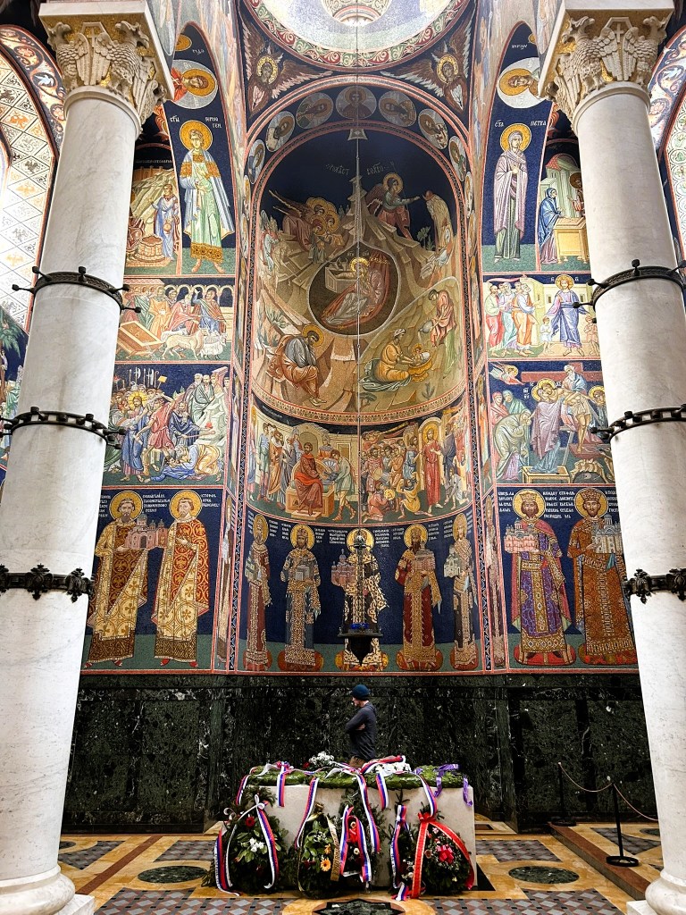 Interior view of a religious building featuring ornate frescoes on the walls, depicting various saints and biblical scenes, with a person standing beside a floral tribute at the base.