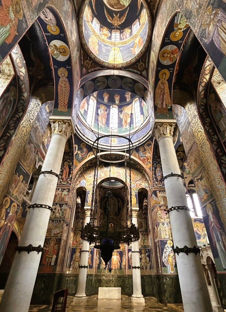 Interior view of a historic church featuring ornate frescoes on the walls and ceiling, tall columns, and a decorative chandelier.
