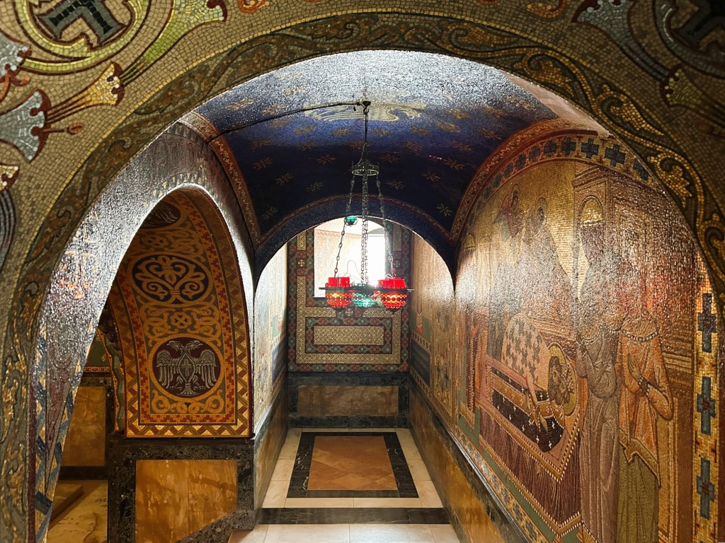 A beautifully adorned corridor featuring intricate mosaics and decorative patterns on the walls and ceiling, illuminated by a red chandelier.