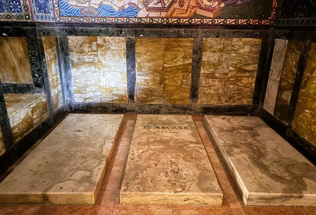 Interior view of a crypt featuring three stone slabs with inscriptions, surrounded by ornate marble walls and decorative mosaics.