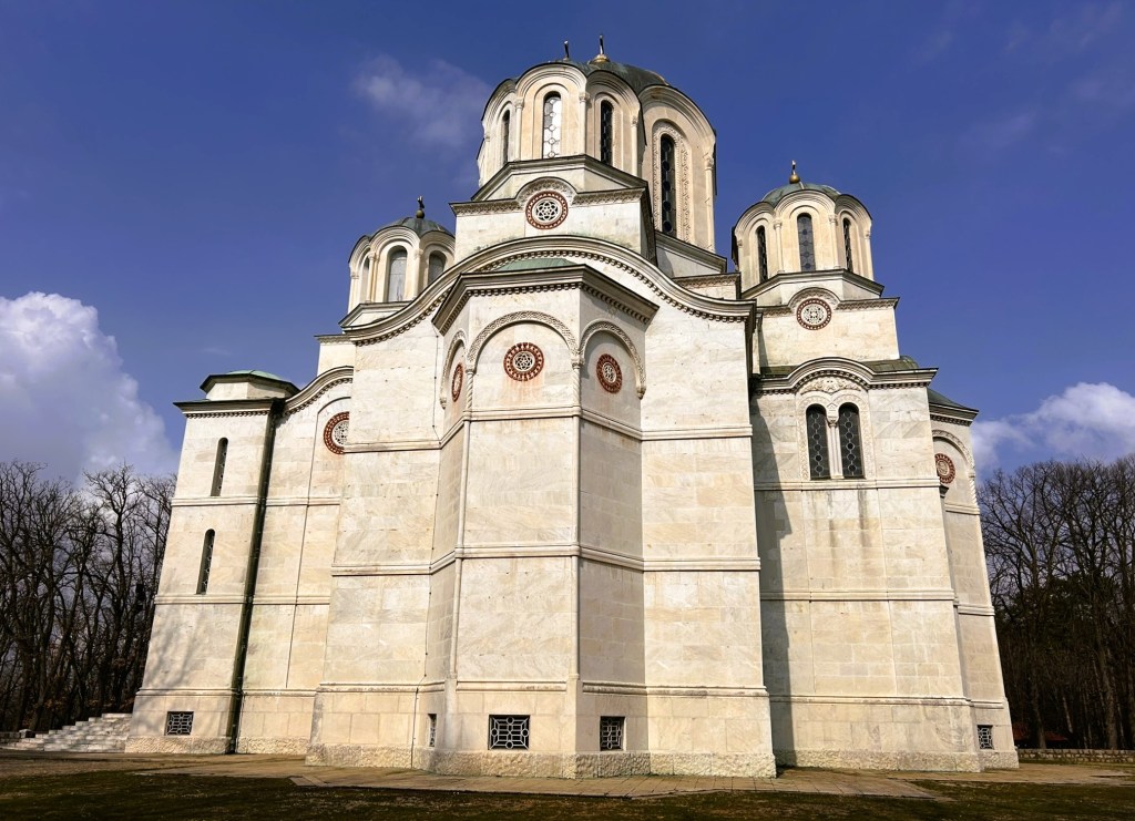 A large, ornate church building with a multi-domed roof, featuring decorative stonework and large windows, set against a blue sky with clouds.