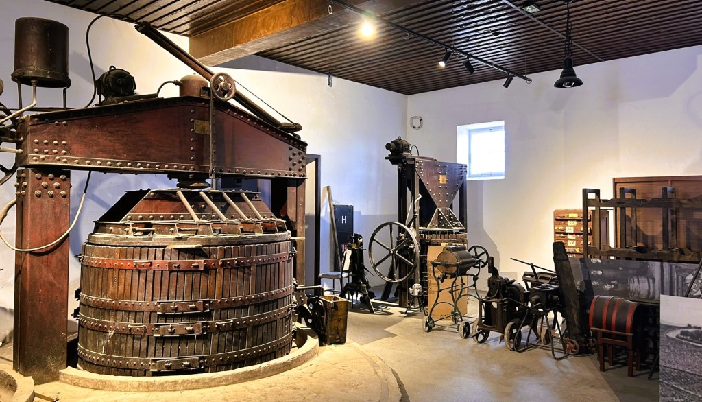 A vintage wine press made of wood and metal, surrounded by various historical agricultural tools and machinery in a museum setting.