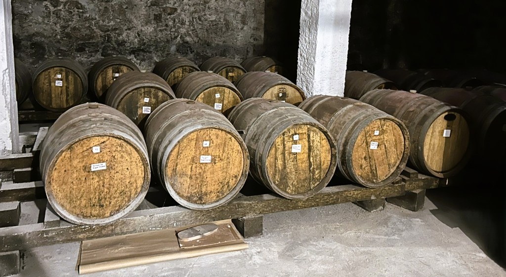 Rows of wooden barrels stacked in a dimly lit cellar, each labelled with a tag, resting on a wooden pallet.