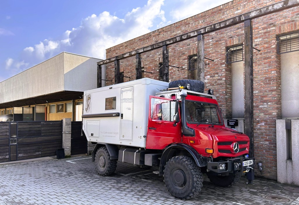 A red Mercedes truck with a white camper unit parked next to a brick building under a partly cloudy sky.