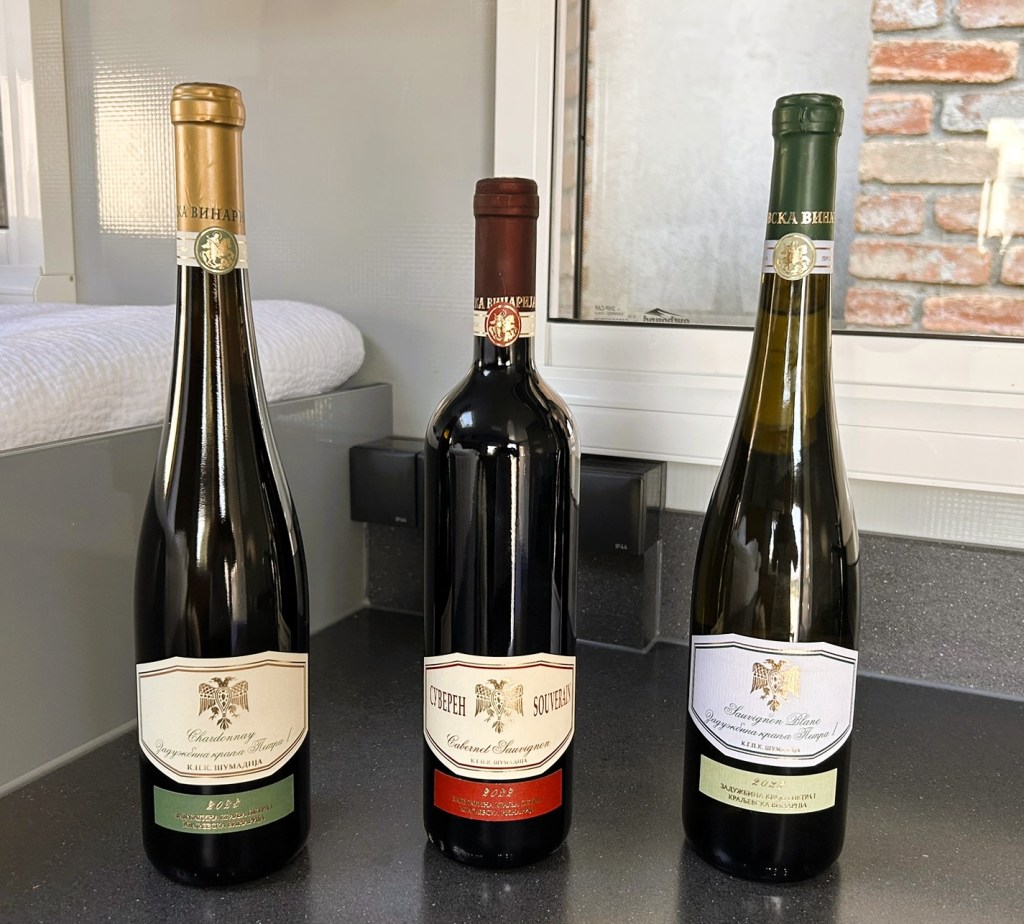 Three bottles of wine on a countertop with a light grey background. The left bottle has a gold cap, the middle bottle has a red cap, and the right bottle has a green cap. Each bottle features a label with different designs and text in various colours.
