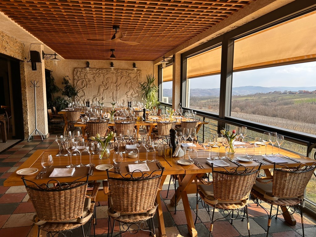A beautifully set dining room with wooden tables and wicker chairs, adorned with glassware and cutlery. There are decorative plants and a stone relief on the wall, with large windows offering a view of a scenic landscape.