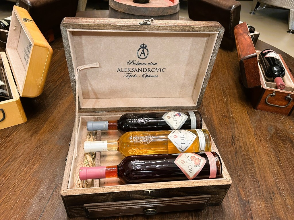 A wooden wine box containing three bottles of wine with labels, resting on a dark wooden surface. Other wine boxes are partially visible in the background.