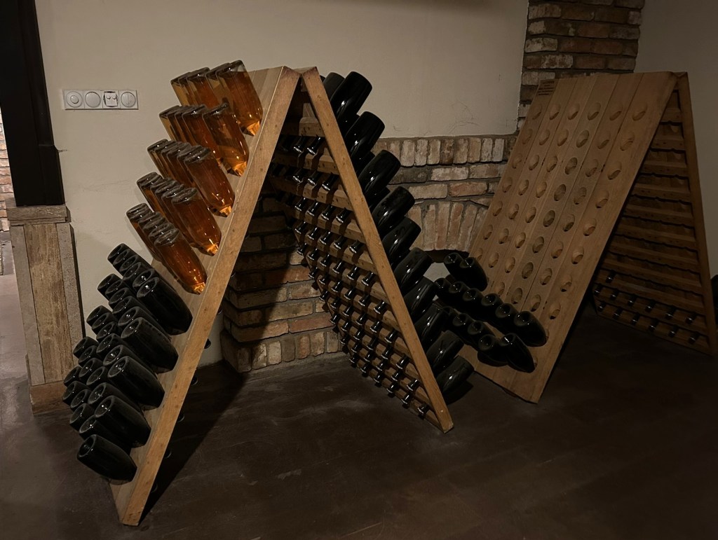 Two wooden racks displaying bottles of sparkling wine, with one rack holding amber bottles and the other black bottles, set against a rustic brick wall.