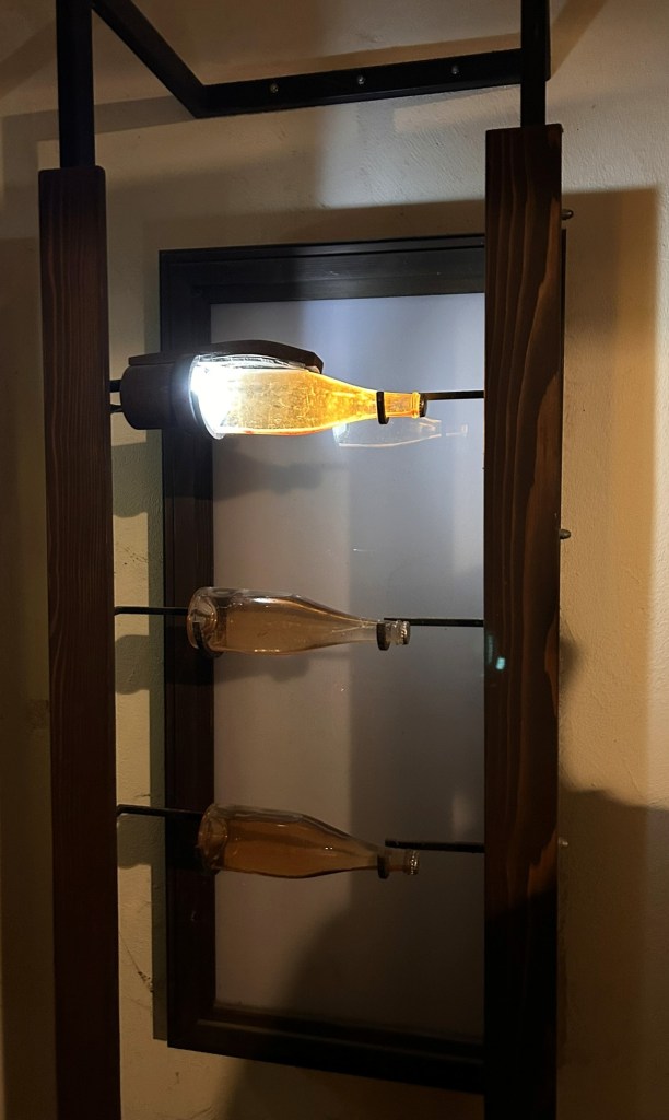 A decorative light fixture featuring a glass bottle illuminated by a bulb, mounted on a wooden frame with two additional bottles hanging below.