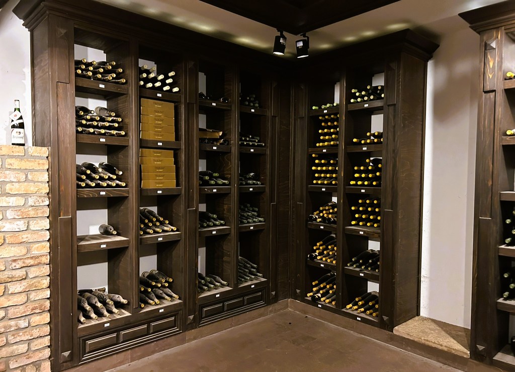 A neatly arranged wine cellar featuring dark wooden shelves filled with various bottles of wine, some with yellow and black labels. There is a brick wall on the left, and the overall atmosphere is warm and inviting.