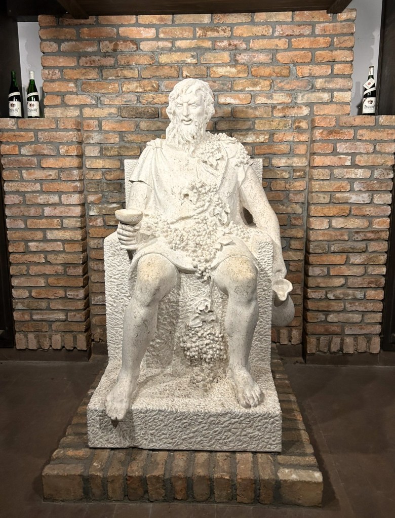 A white stone sculpture of a seated figure with a beard, holding a cup and adorned with grapevines, placed against a brick wall.