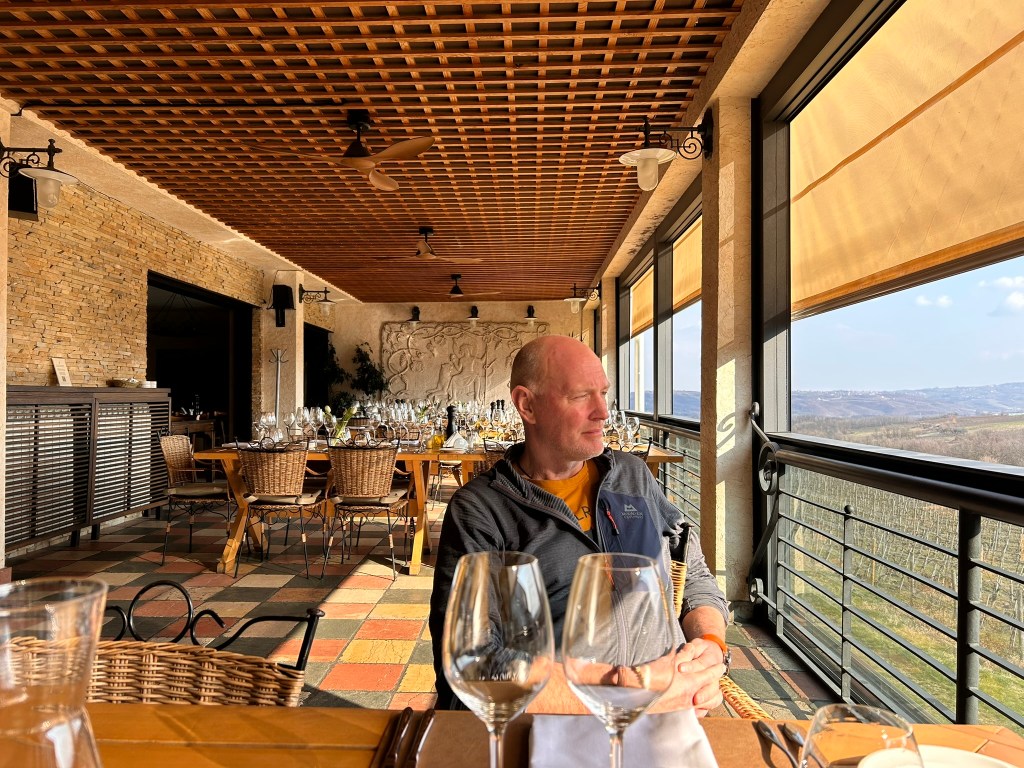 A man sitting at a table with wine glasses, gazing out of a large window in a restaurant with a scenic view of hills.
