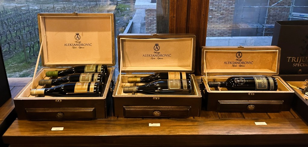 Three wooden wine boxes displaying a selection of Aleksandrović wines, each box containing multiple wine bottles, set against a vineyard backdrop.