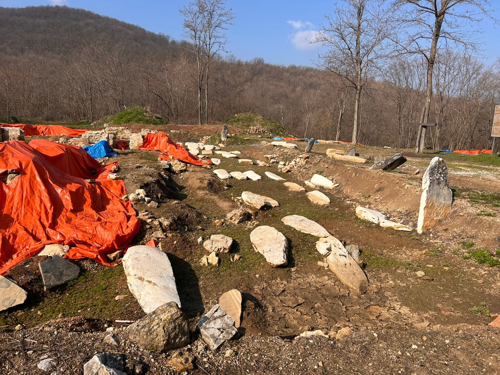 Archaeological site with large stones and orange tarps in a wooded area, featuring a background of bare trees and hills under a blue sky.