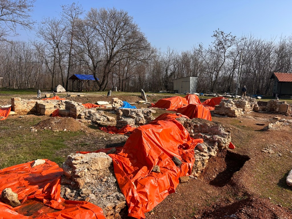 Archaeological site with excavated ruins covered by orange tarps, surrounded by bare trees and a blue shelter in the background.
