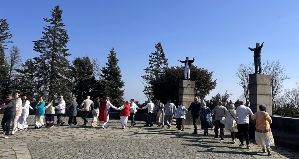 A group of people engaged in a traditional dance around a stone plaza, flanked by two statues on pedestals. The scene features trees in the background and a clear blue sky.