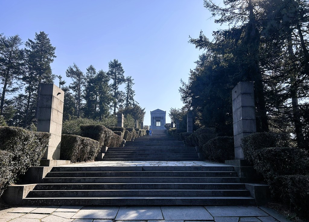 View of a stone staircase leading up to a temple-like structure, surrounded by tall trees and trimmed hedges under clear blue skies.