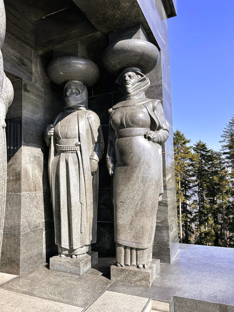 Two large, stylised stone statues of women in traditional attire, each wearing oversized hats, standing against a dark stone wall with trees in the background.